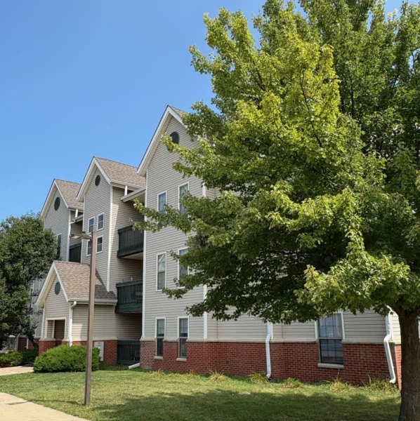 Three-story apartment building with beige siding, red brick lower level, balconies, and several green trees in front under a clear blue sky.