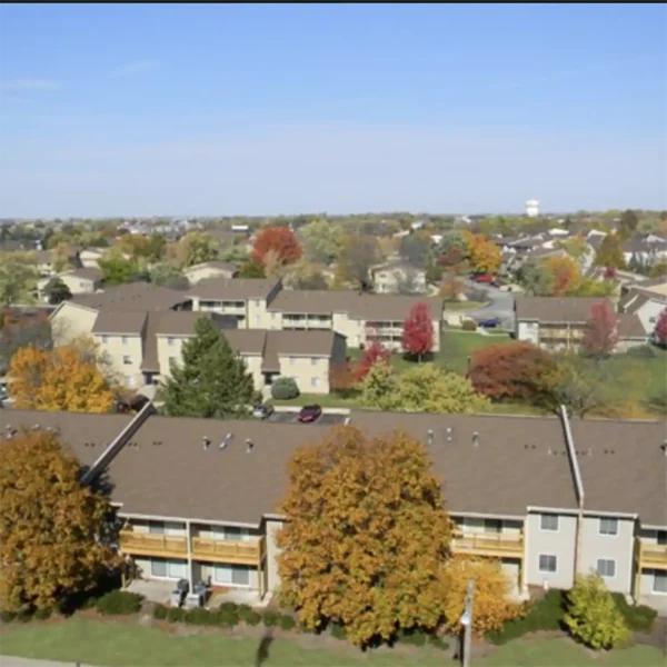 Aerial view of a suburban neighborhood with several tan apartment buildings, green lawns, trees with autumn foliage, and a clear sky.