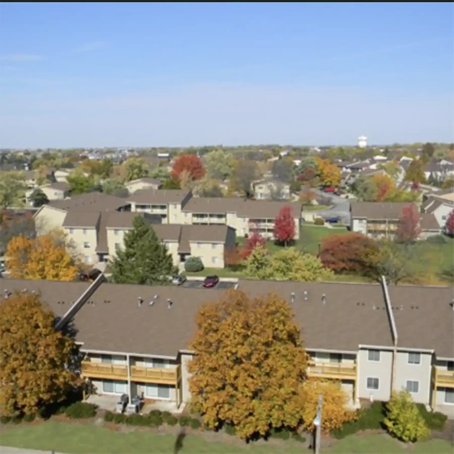 Aerial view of a suburban neighborhood with several tan apartment buildings, green lawns, trees with autumn foliage, and a clear sky.