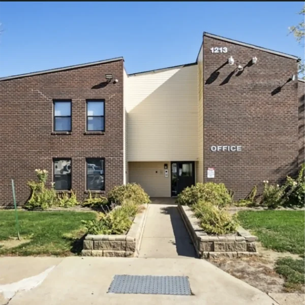 A two-story brick office building with a beige central entrance, labeled "OFFICE" and "1213," surrounded by grass and shrubs under a clear blue sky.
