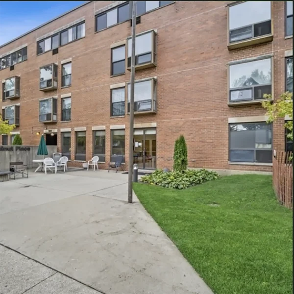 A four-story brick apartment building with multiple windows, a small green lawn, outdoor seating area with chairs and tables, and a paved walkway in front.