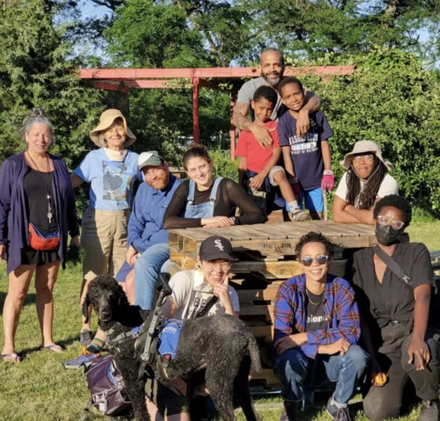 A diverse group of people and a service dog pose together outdoors on a sunny day, standing and sitting around stacked wooden pallets.