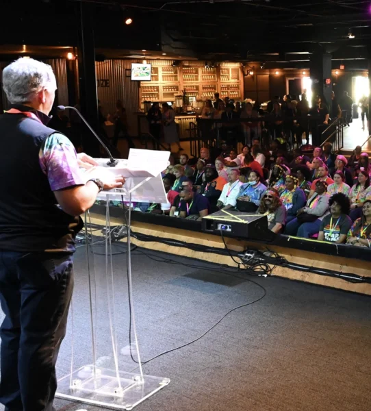 A speaker stands at a clear podium addressing a seated audience in a large indoor venue with a bar in the background.