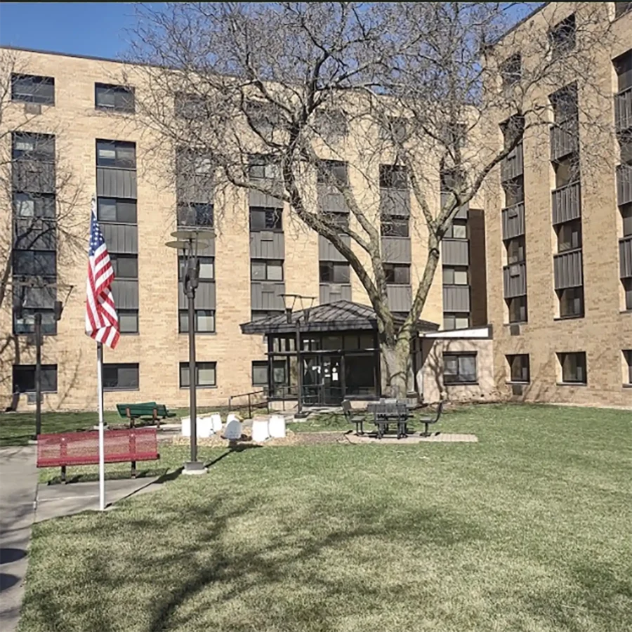 A four-story brick apartment building with a central entrance, surrounded by grass, benches, a flagpole with the American flag, and leafless trees.