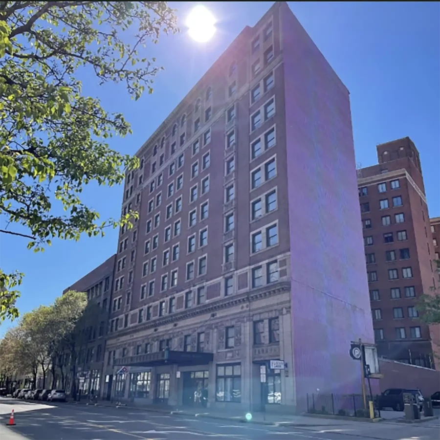 A tall brick building with multiple windows is shown on a sunny day, with sunlight visible above the structure and trees lining the street.