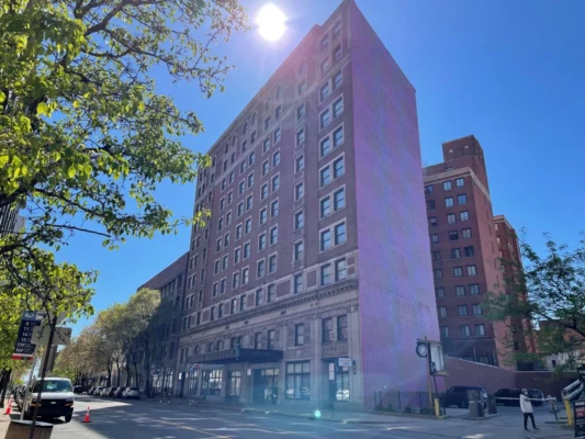 A tall brick building with multiple floors stands on a city street under a clear blue sky with sunlight above. Trees and a few vehicles line the street.