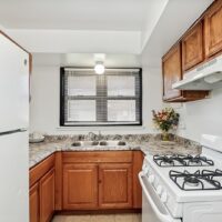 A small kitchen with wooden cabinets, a marble-patterned countertop, a double sink under a window, a white refrigerator, and a white gas stove. A vase of flowers sits on the counter.