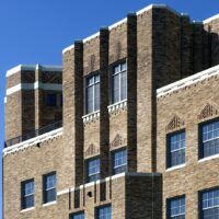 A multi-story brick building features geometric Art Deco architectural details, with tall vertical window arrangements and decorative patterns, set against a clear blue sky.