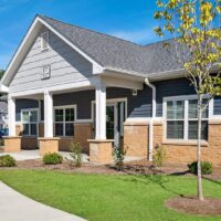 Single-story residential building with gray siding, tan stone accents, white columns, and a front porch, surrounded by a manicured lawn and small shrubs under a blue sky.