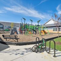 A playground with green and orange climbing equipment, a bench, a bike rack with two bikes, and surrounding buildings under a blue sky.