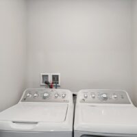 A white washing machine and dryer set side by side in a laundry area with plain white walls and visible water hookups behind them.