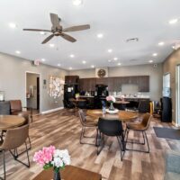 A modern communal kitchen and dining area with round tables, brown chairs, a ceiling fan, and a bulletin board on the wall. The space is well-lit and decorated with flowers.