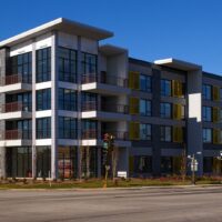 A modern three-story apartment building with large windows, balconies, and yellow accents, situated on a street corner under a clear blue sky.