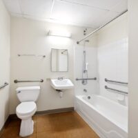 Accessible bathroom with grab bars, a toilet, wall-mounted sink, mirror, towel racks, and a bathtub-shower combination with handheld showerhead.