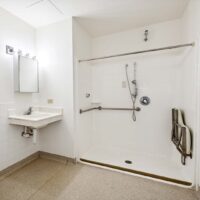 Accessible bathroom with a roll-in shower, grab bars, handheld showerhead, shower seat, wall-mounted sink, and a mirror with overhead lighting.