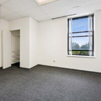 Empty office room with gray carpet, white walls, an open closet with shelves, a window with partial vertical blinds, and a view of trees outside.