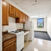 Small kitchen with wooden cabinets, white stove, refrigerator, sink, and a window in the background letting in natural light.