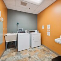 Laundry room with an orange and gray wall, a washing machine, a dryer, a utility sink, and various instructional signs on the walls.