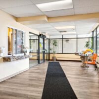 A clean and bright office lobby with a reception window, glass walls, orange chairs, a black floor mat, and potted plants.