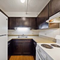 Small L-shaped kitchen with white appliances, dark brown cabinets, light countertops, stainless steel sink, and wood flooring under overhead lighting.
