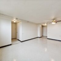 Empty room with white walls, tiled floor, ceiling lights, ceiling fan, and visible doorways leading to other areas.