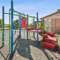 A colorful playground structure with blue poles, green bars, and red slides and stairs, set on a gravel surface near a tan building and a chain-link fence.