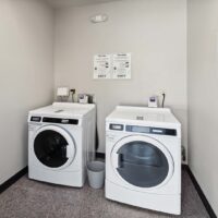 A laundry room with a front-load washing machine and dryer side by side, a trash bin between them, and service instructions posted on the wall above.