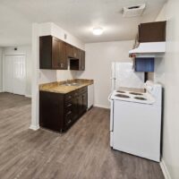 Small kitchen with dark cabinets, tan countertop, white stove, and white refrigerator. Wood-style flooring extends into the adjacent room. Walls are off-white.