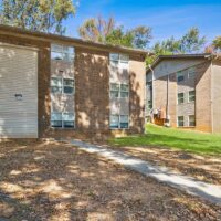 Two brick apartment buildings with multiple windows, a sloped grassy area, a sidewalk, and scattered autumn leaves under a clear blue sky.