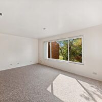 Empty room with light-colored walls and carpeted floor, featuring a large window with a view of trees and a brick wall outside.
