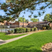 A fenced outdoor pool area with a covered patio, surrounded by green lawns, trees, and brick buildings under a clear blue sky.