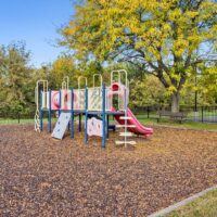 A playground with climbing features, slides, and ladders stands on a surface covered with mulch, surrounded by trees and a metal fence.