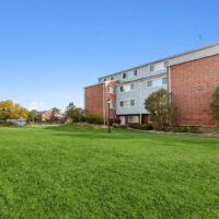 A three-story brick and siding apartment building stands behind a large, well-maintained grassy area under a clear blue sky.