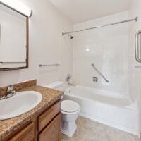 A bathroom with a sink, mirror, toilet, bathtub-shower combo, white tile walls, and brown countertop on a wooden vanity.