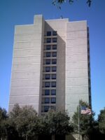 A tall, rectangular concrete building with multiple windows stands behind trees; a U.S. flag is visible in the foreground.
