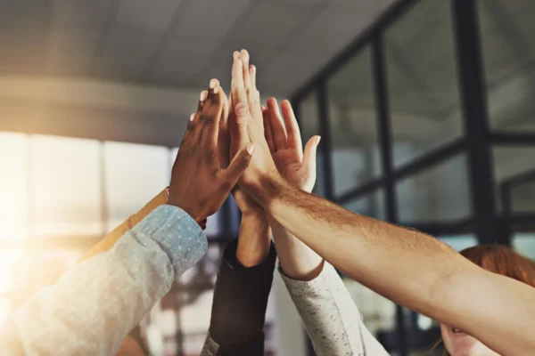 Four people standing in an office environment put their hands together for a high-five, symbolizing teamwork and unity.