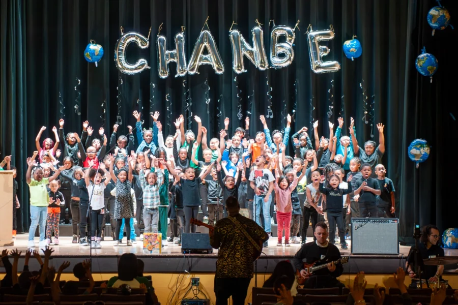 A group of children perform on stage under balloon letters spelling "CHANGE," with musicians playing in front and decorative globe balloons on the sides.