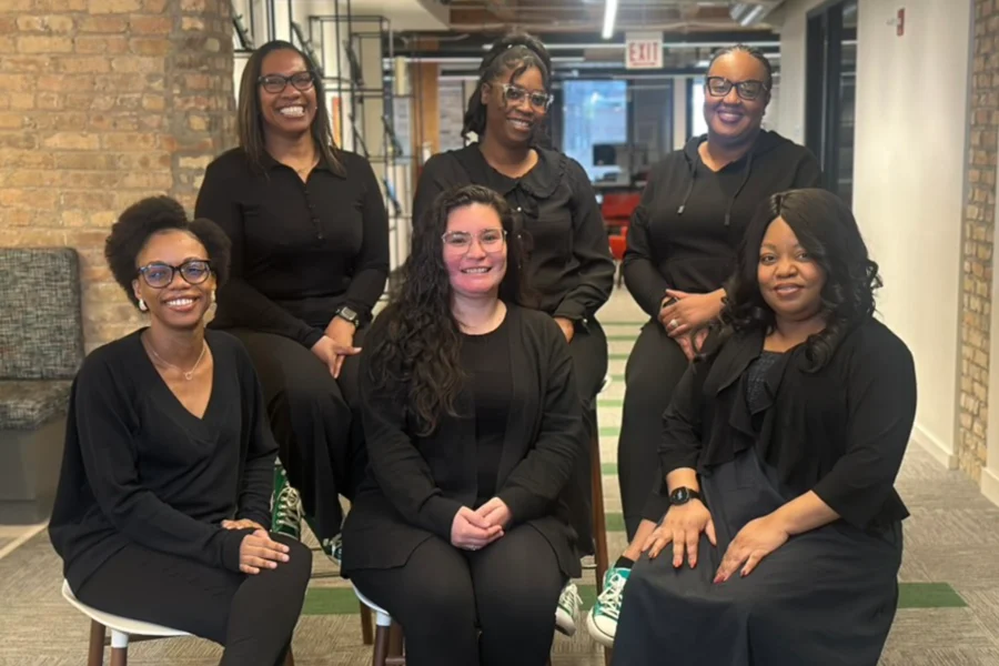 Six women dressed in black sit and stand in an office space, smiling at the camera. Exposed brick and modern office furniture are visible in the background.