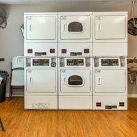 Three stacked commercial washer and dryer units with digital controls are lined up against a wall in a laundry room with wood flooring.