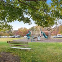 A park scene with a bench in the foreground, a playground with slides and climbing structures in the middle, and parked cars in the background among trees.