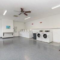 A clean laundry room with five top-loading washing machines, two front-loading dryers, a wall-mounted table, and a ceiling fan.