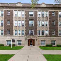A four-story brick apartment building with decorative stonework, symmetrical windows, and a central arched entrance, surrounded by green lawns and a concrete walkway.