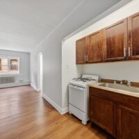 Small kitchen area with dark wood cabinets, a white gas stove, and a single sink; adjacent room with wood flooring and window visible through an archway.