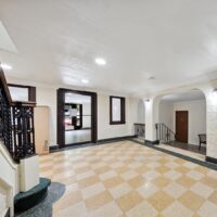 A well-lit building lobby with checkered tile floor, cream walls, a staircase with a black railing, arched doorways, and doors on either side.