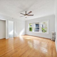 Bright, empty room with hardwood floors, white walls, a ceiling fan, large windows letting in daylight, and a radiator below one window.