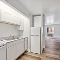 A small kitchen with white cabinets, a sink, a stove, and a white refrigerator, opening into a living area with wood flooring and a window.