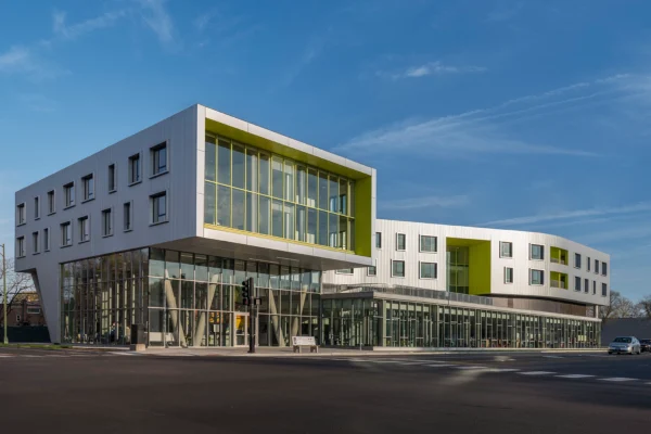Modern, multi-story building with large glass windows, white panels, and green accents, located at a street corner under a clear blue sky.