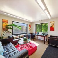 Modern office with black sofas, glass coffee table on a red rug, large windows with outdoor views, desk, and wall art.