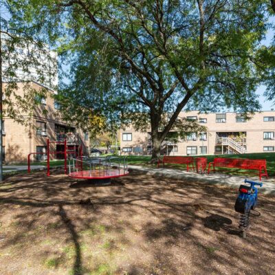 A playground with a merry-go-round, red benches, a spring rider, and climbing bars is surrounded by apartment buildings and a large tree.