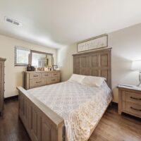 A bedroom with a wooden bed, white patterned bedding, matching dresser and nightstand, a lamp, and a framed sign above the headboard.